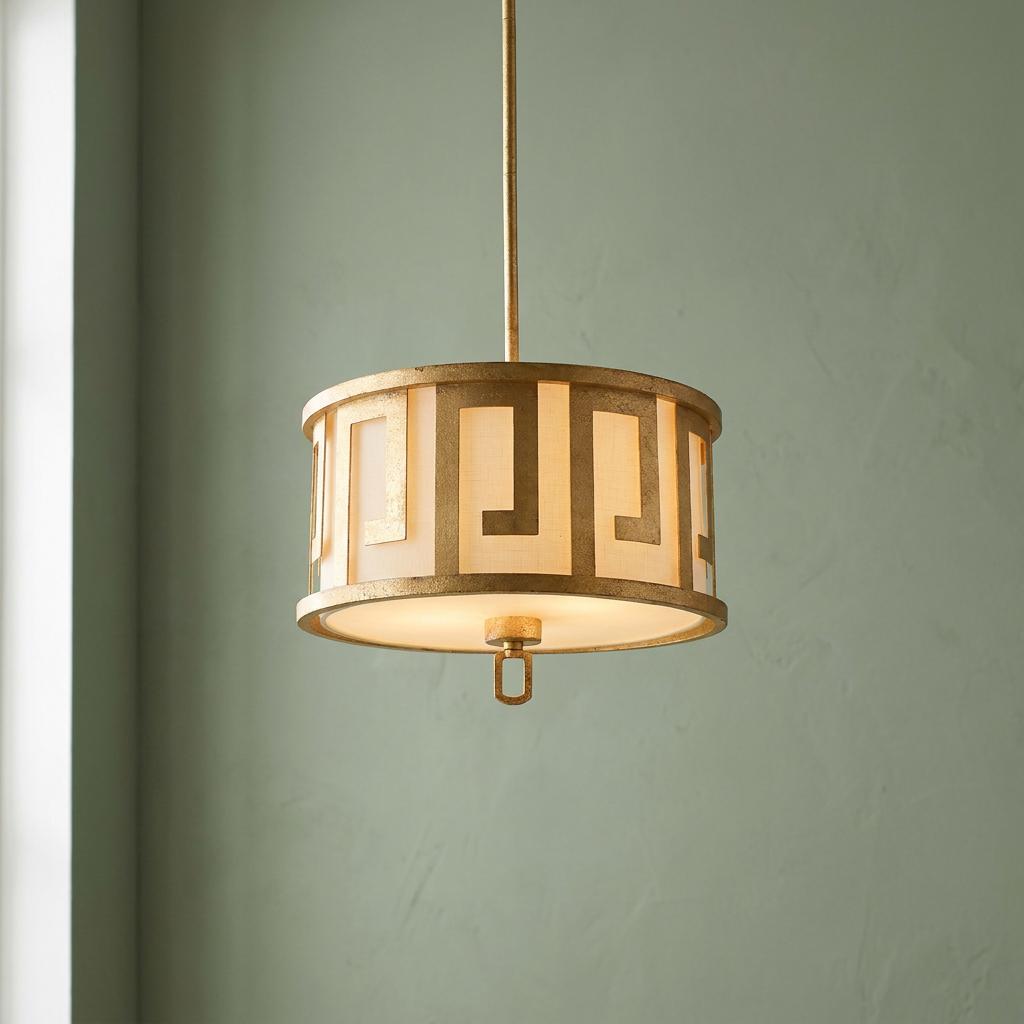 A gilded drum pendant with a Greek key pattern and fabric shade hanging against a muted green wall.