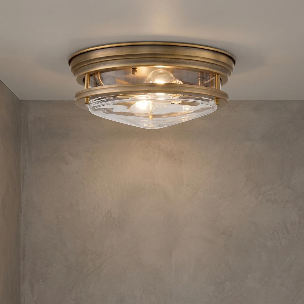 A brushed bronze flush mount bathroom ceiling light with clear glass and two bulbs against a textured wall.