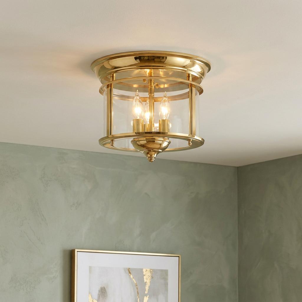 Polished brass flush mount ceiling light with clear glass and three candle bulbs against a green wall.