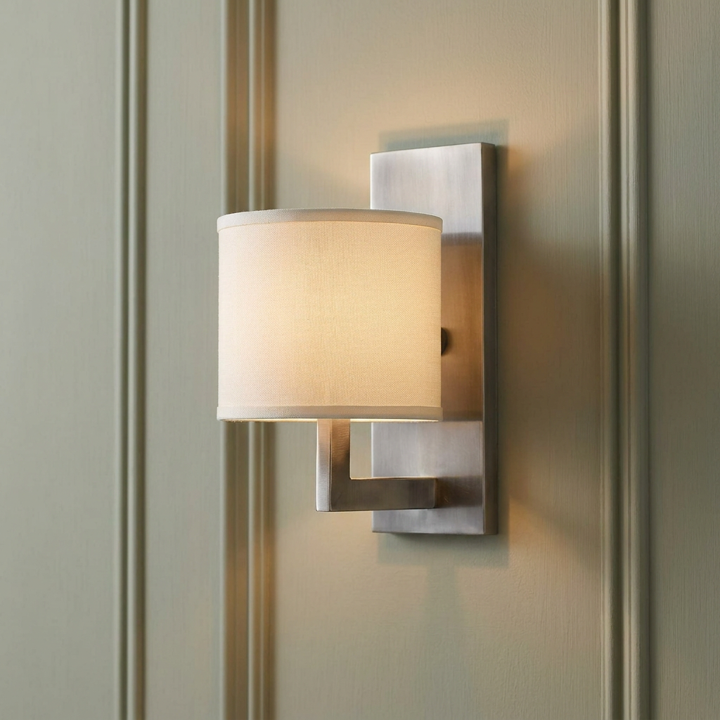 Hinkley Hampton nickel wall light with a white fabric shade, illuminated on a beige paneled wall.