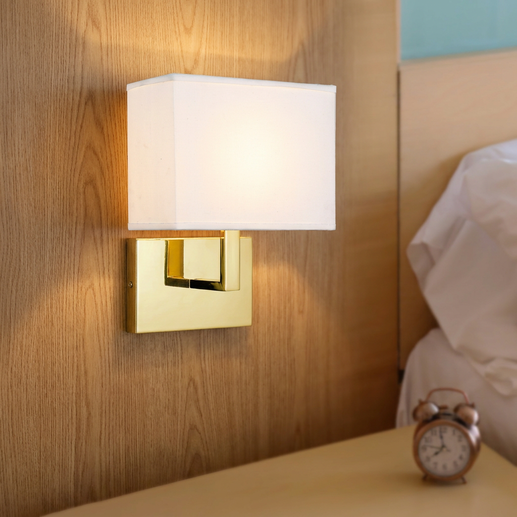 Satin brass wall light with a white fabric shade mounted on a wooden headboard in a bedroom setting.