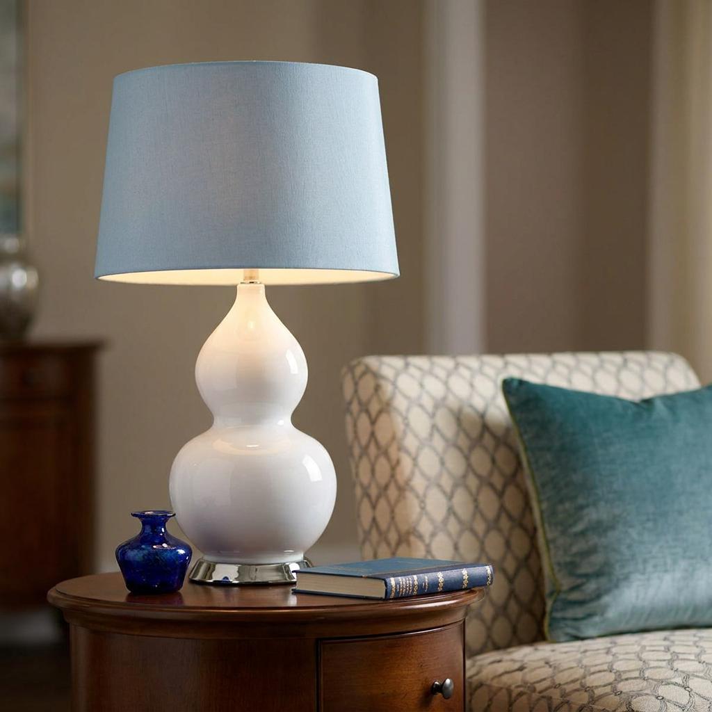 Isla white ceramic table lamp with a light blue shade on a dark wood table beside a patterned armchair.