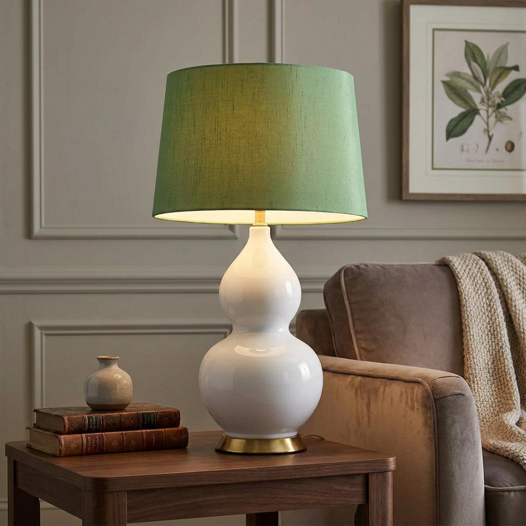 White gourd-shaped ceramic lamp with a green fabric shade on a wooden table in a cozy living room.