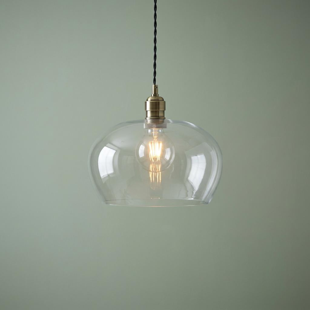 Brass and clear glass pendant light with a lit filament bulb against a soft green background.