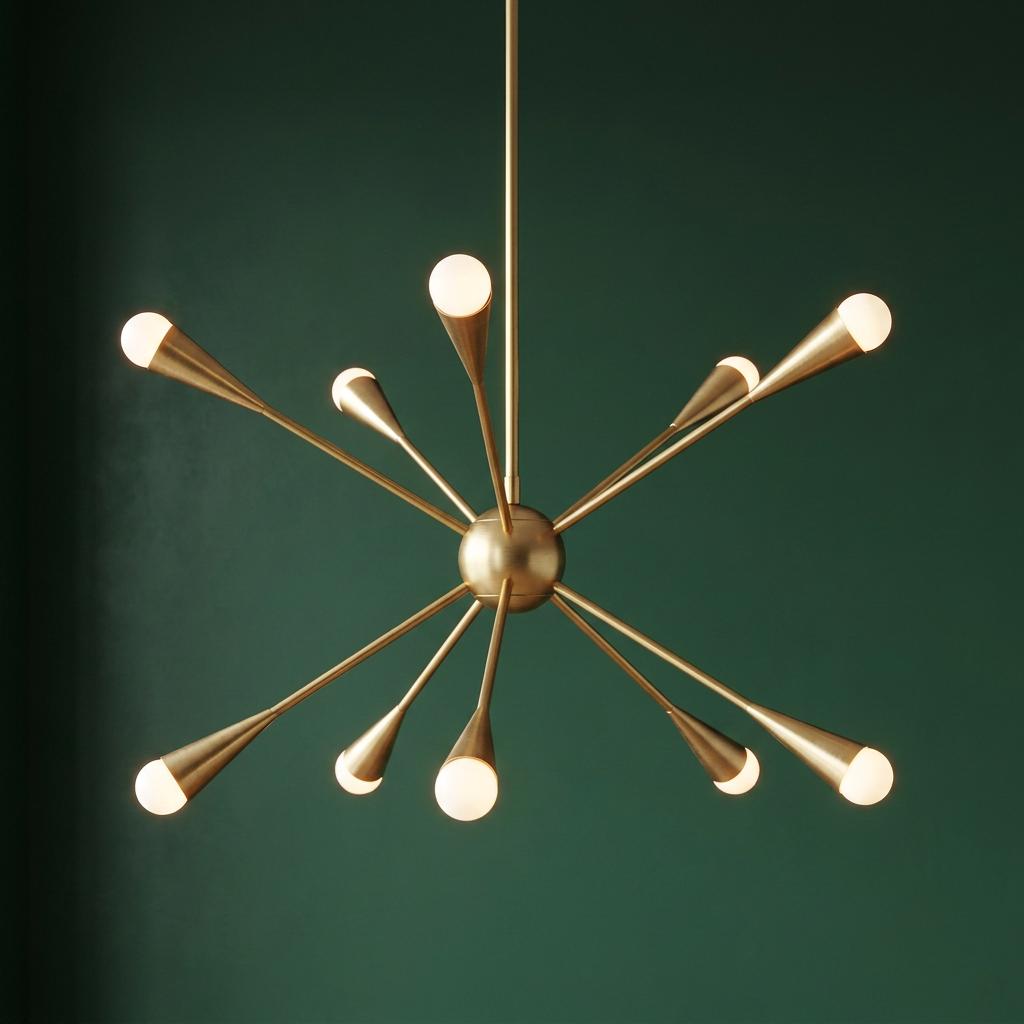 Modern Jax 10 light brass chandelier with a starburst design and orb bulbs against a dark green wall.