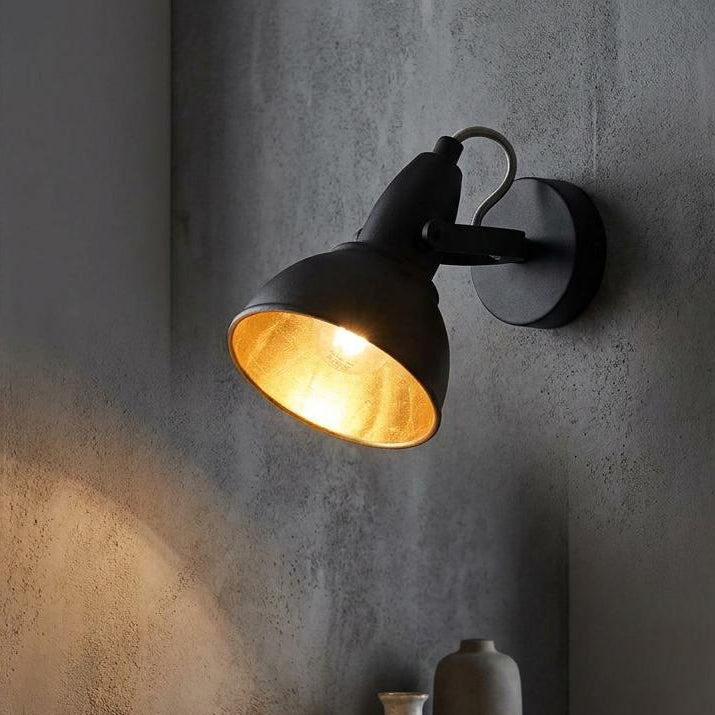 Jurva Urban black adjustable wall spotlight with a gold interior, mounted on a concrete wall above a wooden shelf.