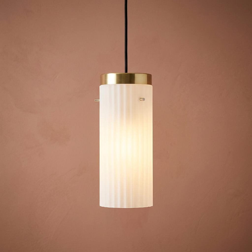 Kansa Bula prismatic fluted glass cylinder pendant light with a brass cap, glowing against a pink textured wall.