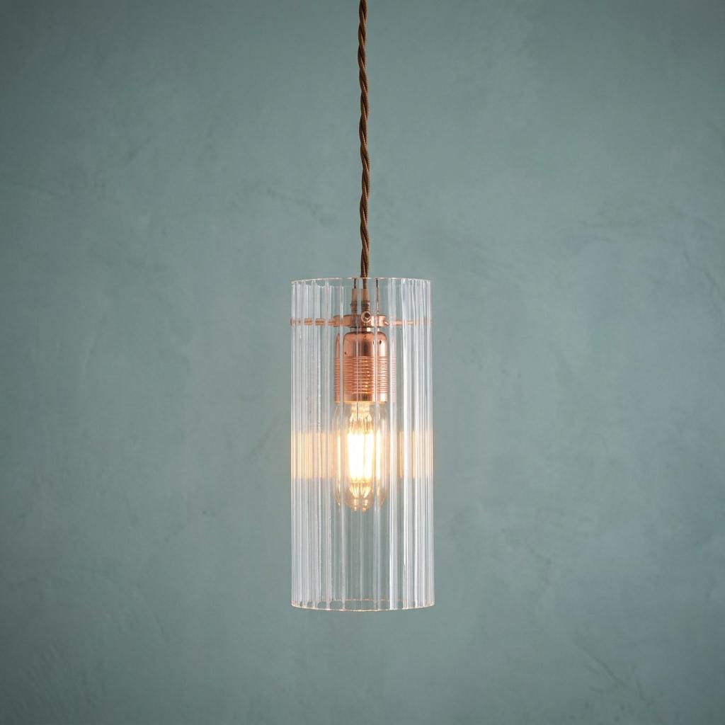 Lit Kansa elongated prismatic ceiling light with copper bulb holder and clear ribbed shade on a textured teal wall.