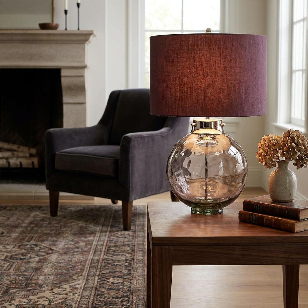 A Kara Polished Nickel and Smoke Glass Table Lamp with a plum drum shade on a wooden side table.