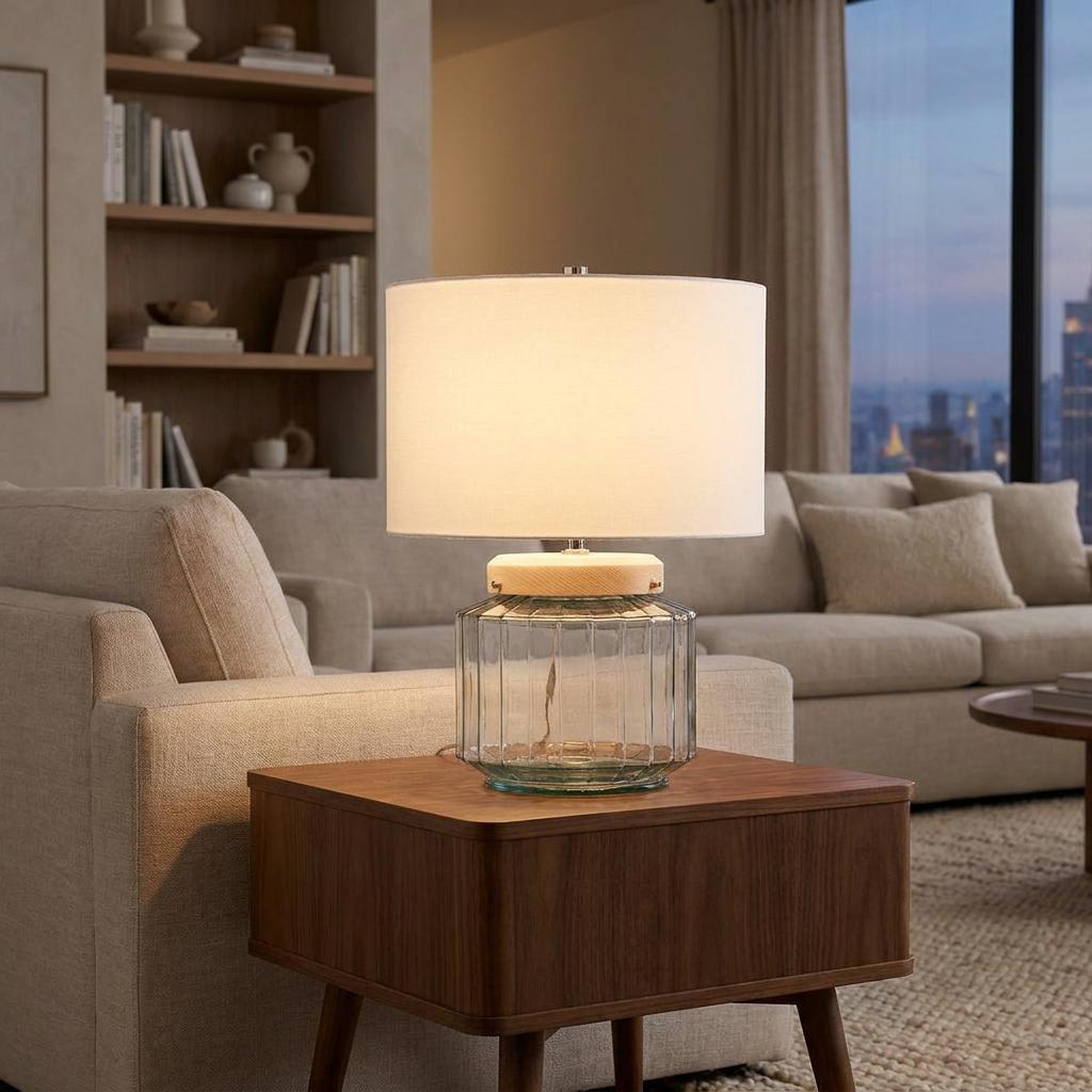 Illuminated Luga glass table lamp with a white drum shade on a wooden side table in a modern living room.