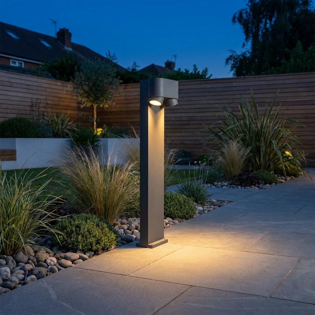 A grey Lutec Cypres bollard light casts a warm glow over a paved garden path in a landscaped backyard at dusk.