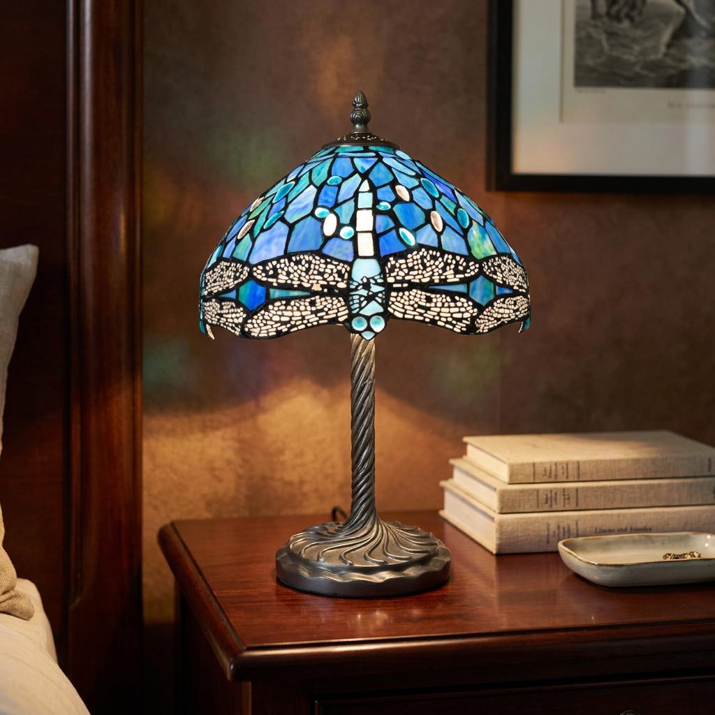 Tiffany-style blue dragonfly table lamp glowing on a wooden bedside table next to a stack of books.