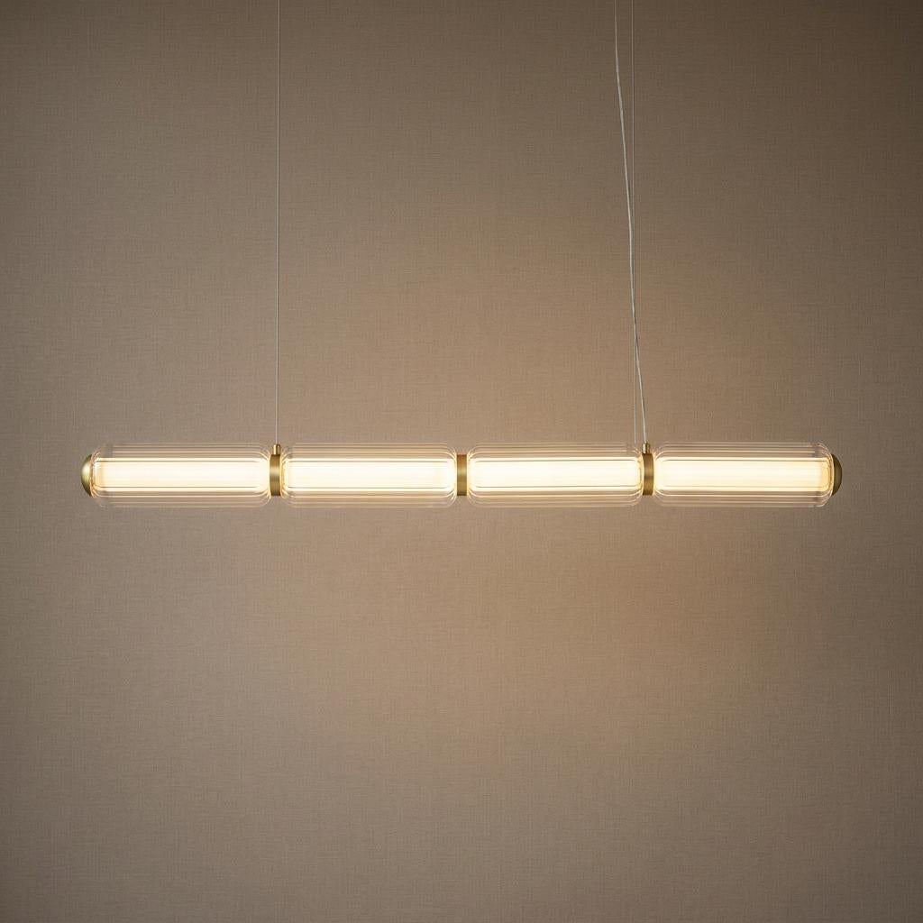 Linear LED pendant with four clear ribbed glass cylinders and satin brass accents against a neutral wall.