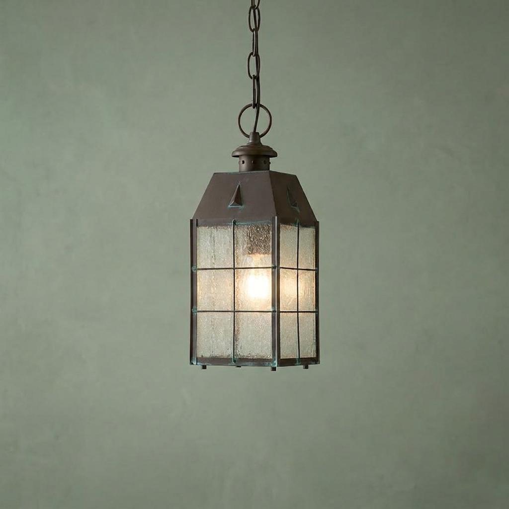 Weathered brass outdoor chain lantern with seeded glass panes hanging against a textured green wall.
