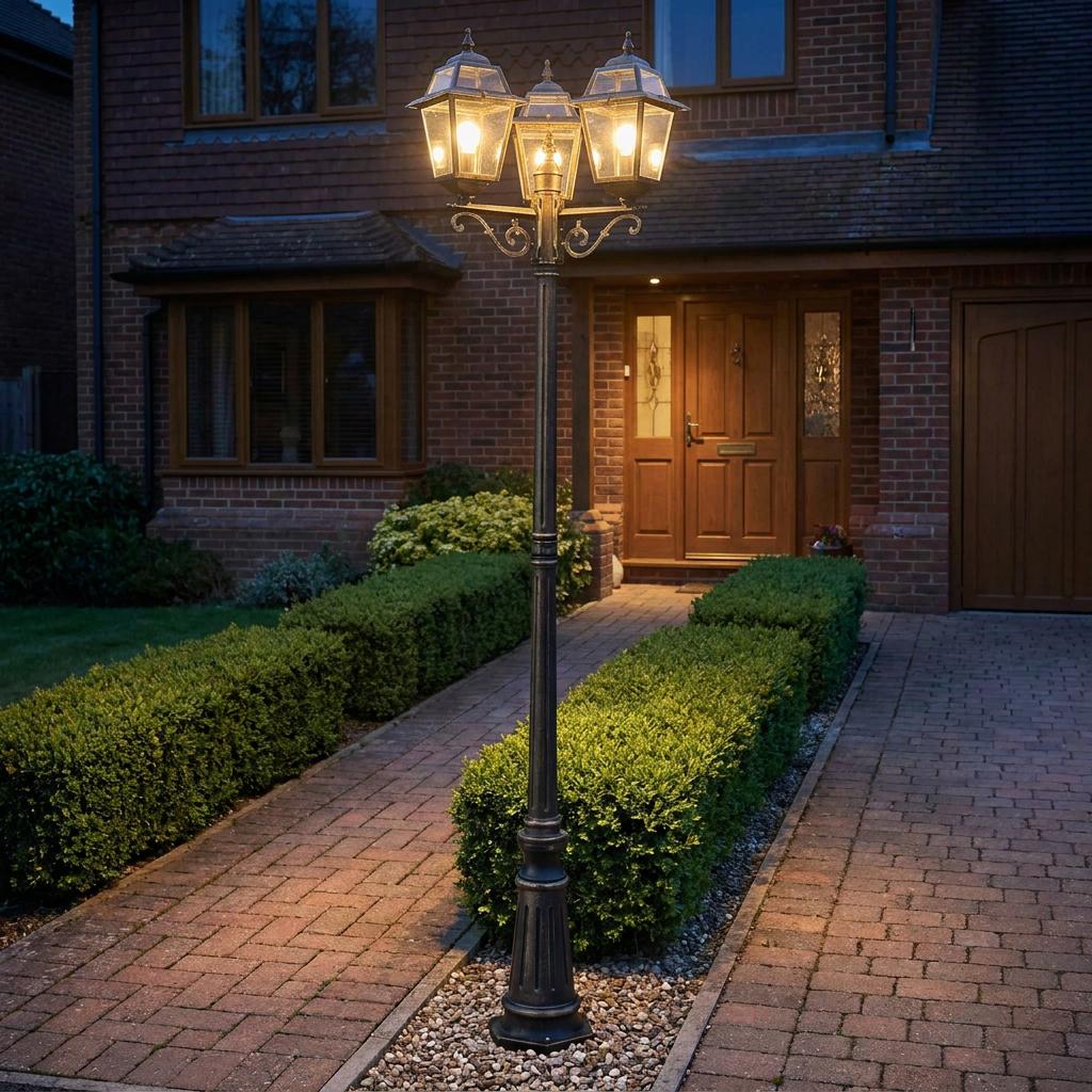 A black and gold three-light outdoor lamp post illuminating a brick path and manicured hedges at night.
