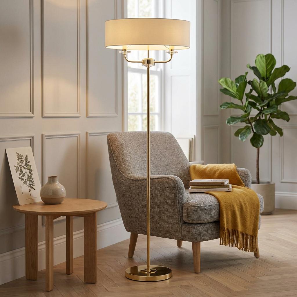Nixon brass floor lamp with a wide cream drum shade standing next to a grey armchair in a paneled room.