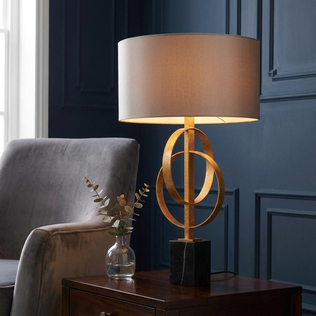 Gold table lamp with interlocking rings and mink shade on a marble base, set on a dark wood table.