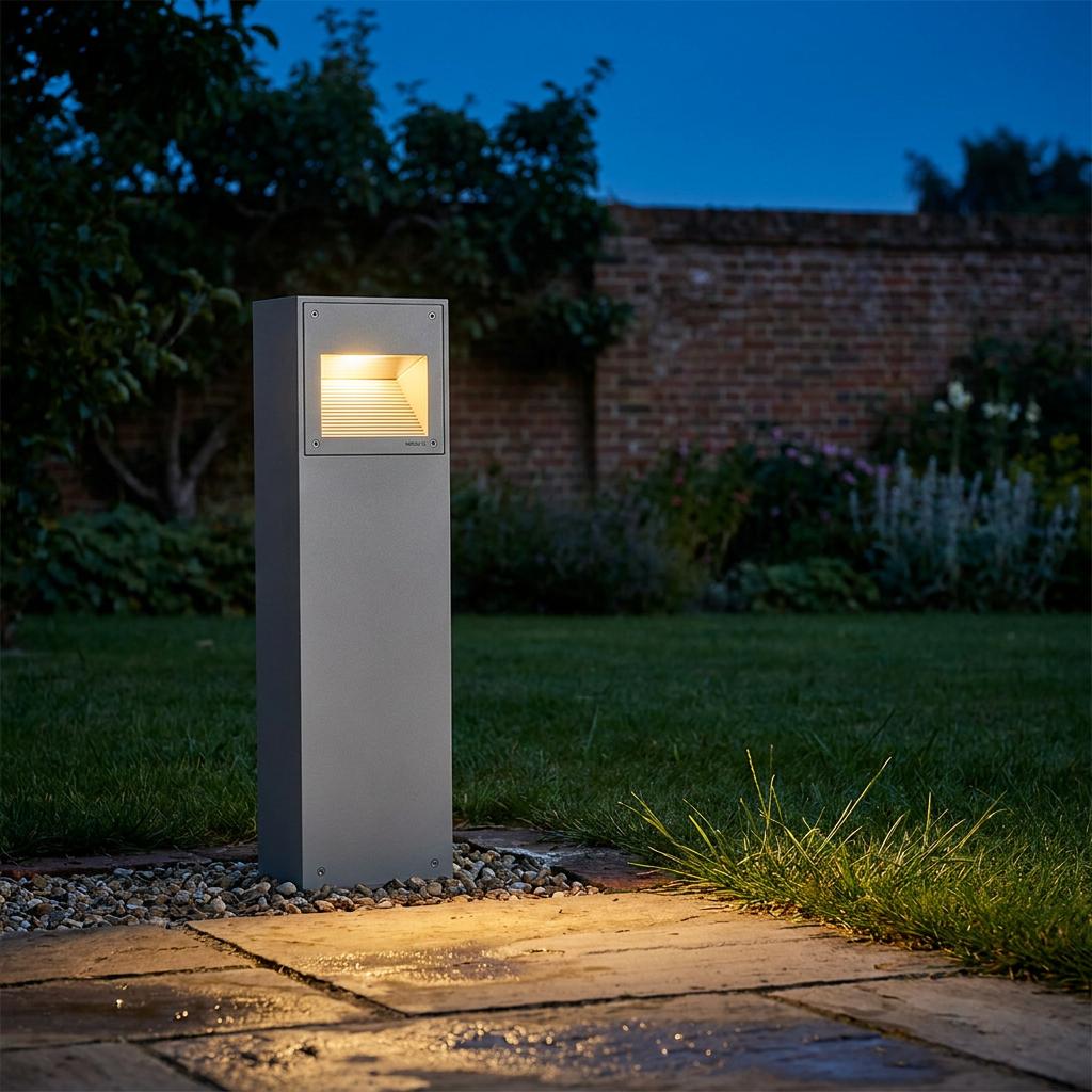 Norlys Namsos tall aluminium outdoor bollard light glowing on a garden path at night near a brick wall.