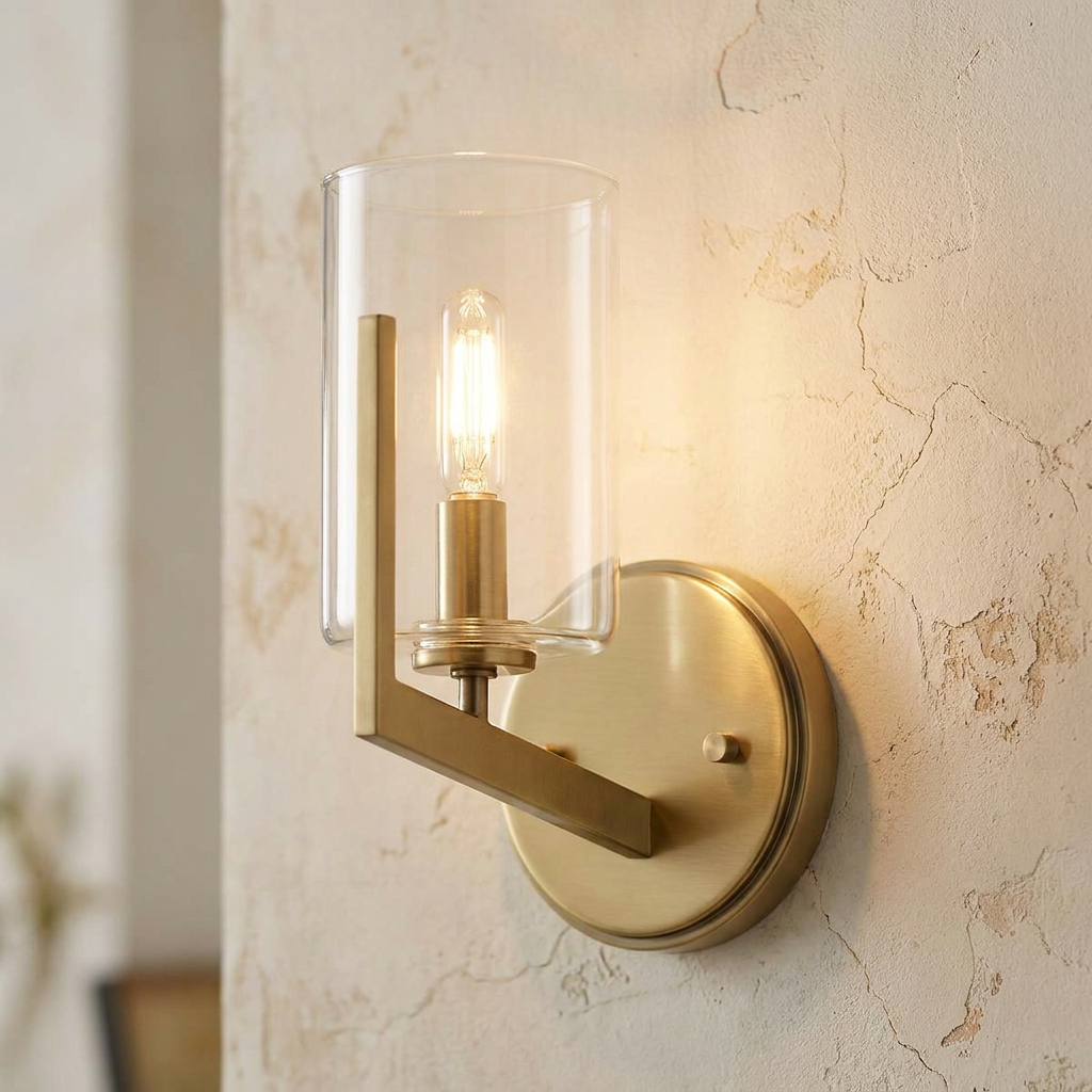 Brass wall sconce with a cylindrical glass shade and warm bulb on a textured, neutral-toned wall.