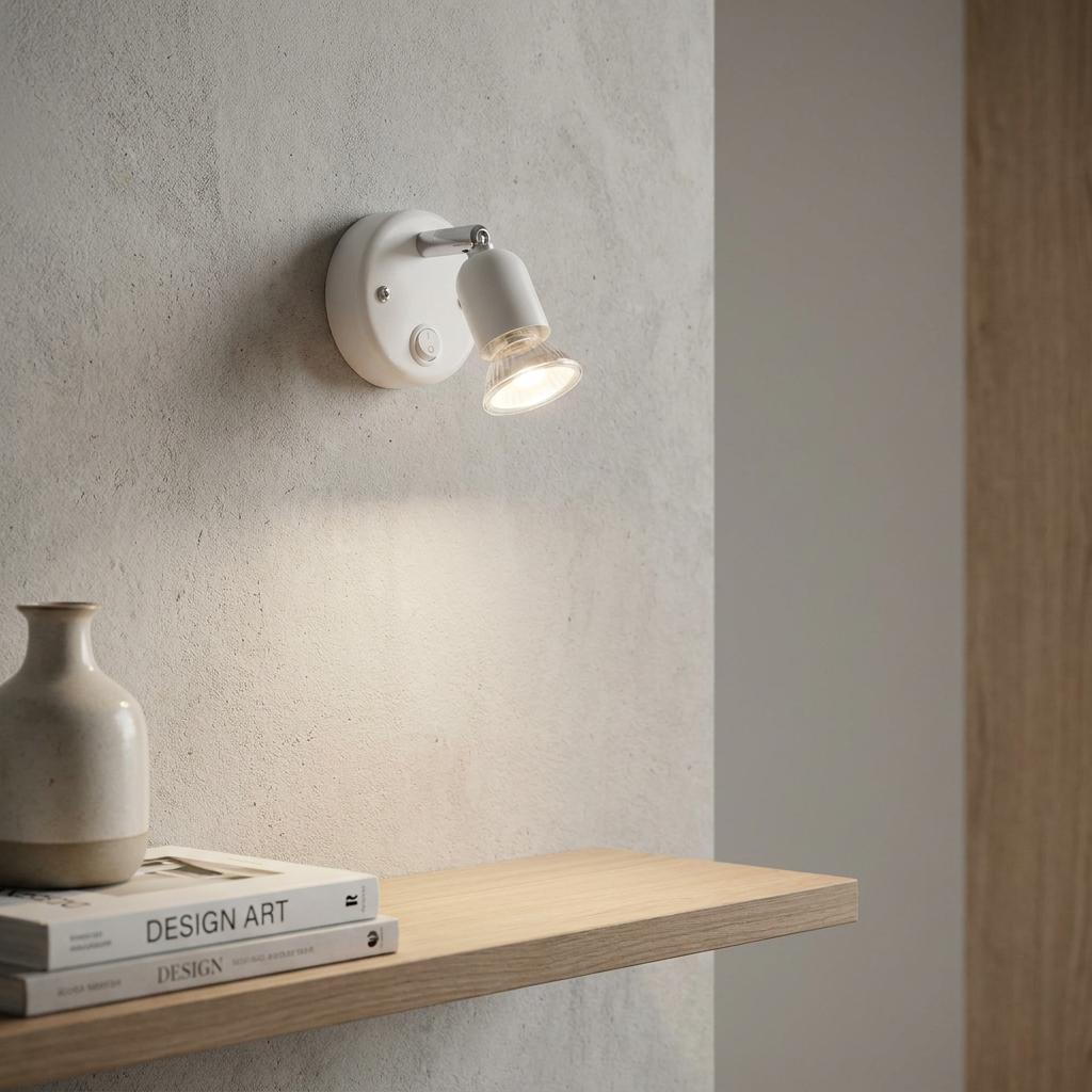 White adjustable switched spotlight mounted on a wall, illuminating a shelf with art books and a vase.