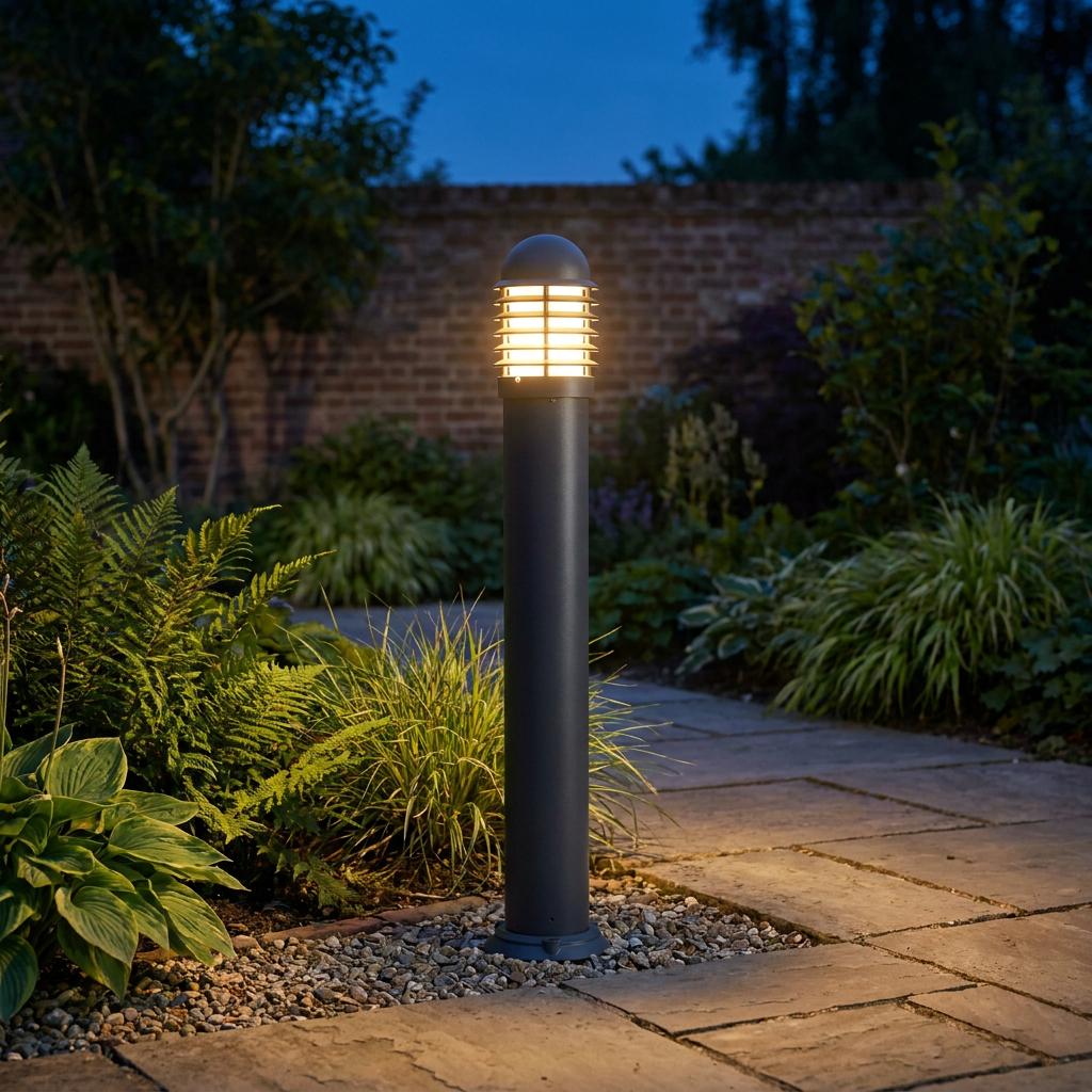 A black outdoor bollard light glowing warmly on a stone garden path at dusk, surrounded by lush green foliage.