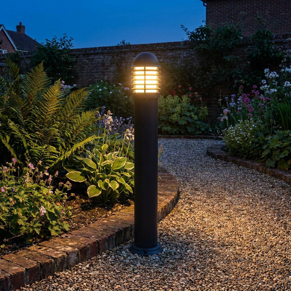Illuminated black bollard light with a cage design standing on a gravel garden path at dusk.