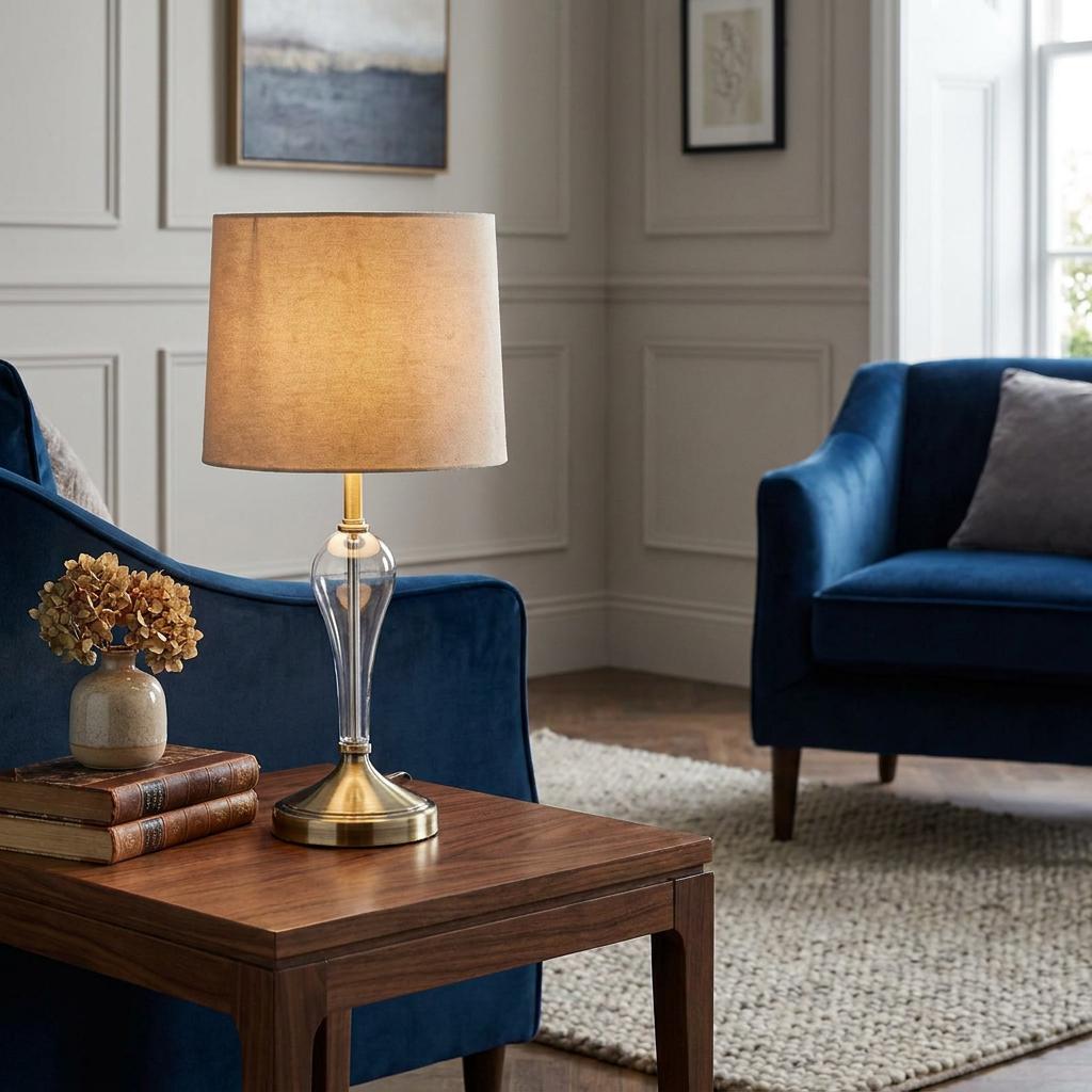 Antique brass table lamp with a glass stem and beige shade on a wooden table next to a blue velvet armchair.