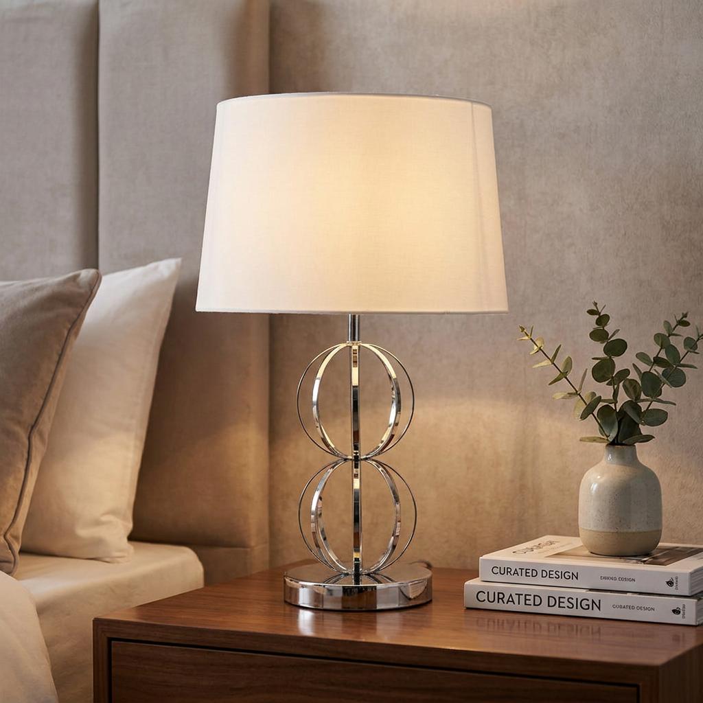 Chrome table lamp with interlocking circles and a white shade on a bedside table.
