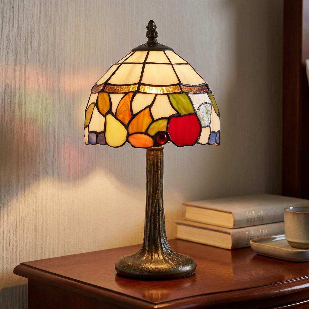 Warmly lit fruit-patterned Tiffany lamp on a bronze base sitting on a wooden nightstand beside stacked books.