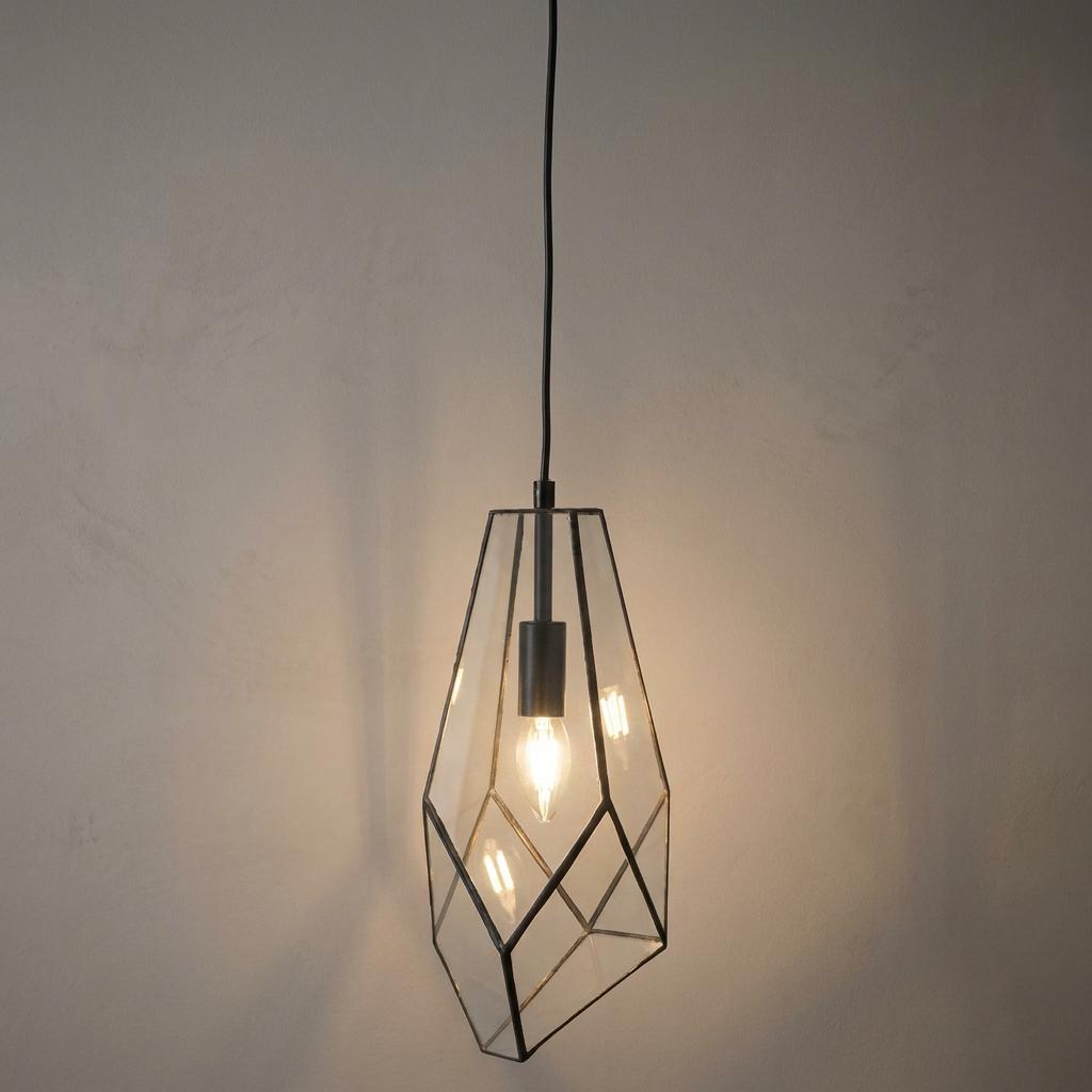 Faceted geometric glass pendant light with a black metal frame and glowing bulb against a neutral wall.