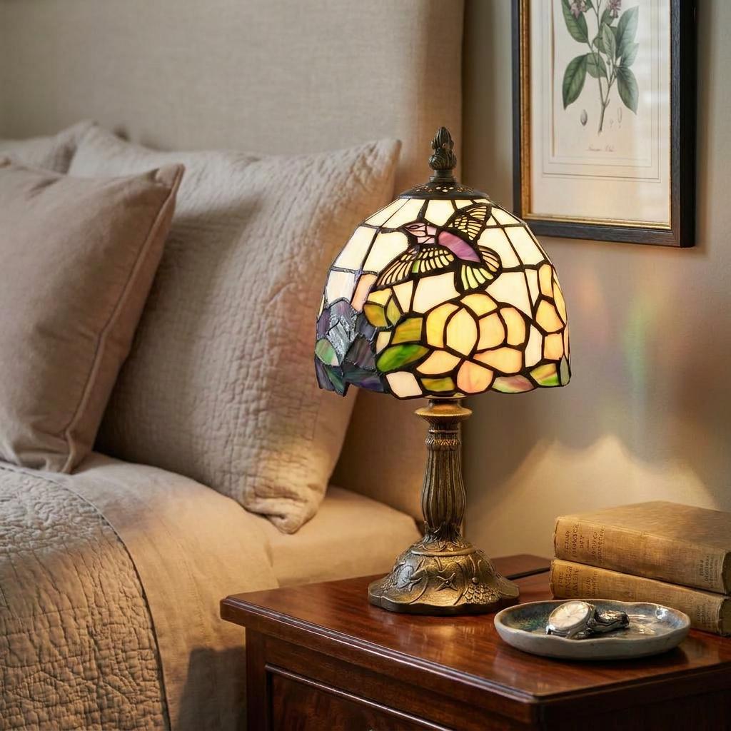 Lit Tiffany hummingbird bedside lamp on a wooden nightstand, featuring a colorful stained glass floral shade.