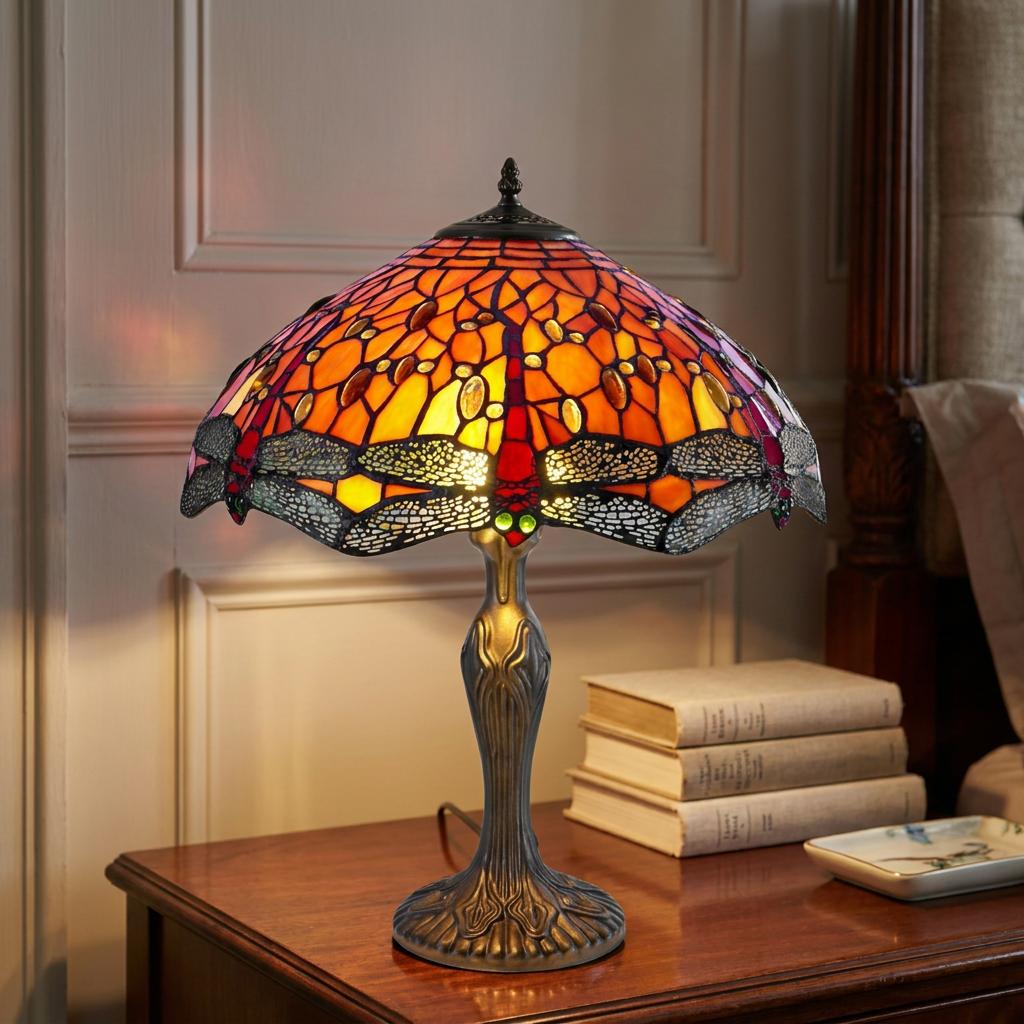 Orange dragonfly Tiffany table lamp glowing on a wooden bedside table next to a stack of books.