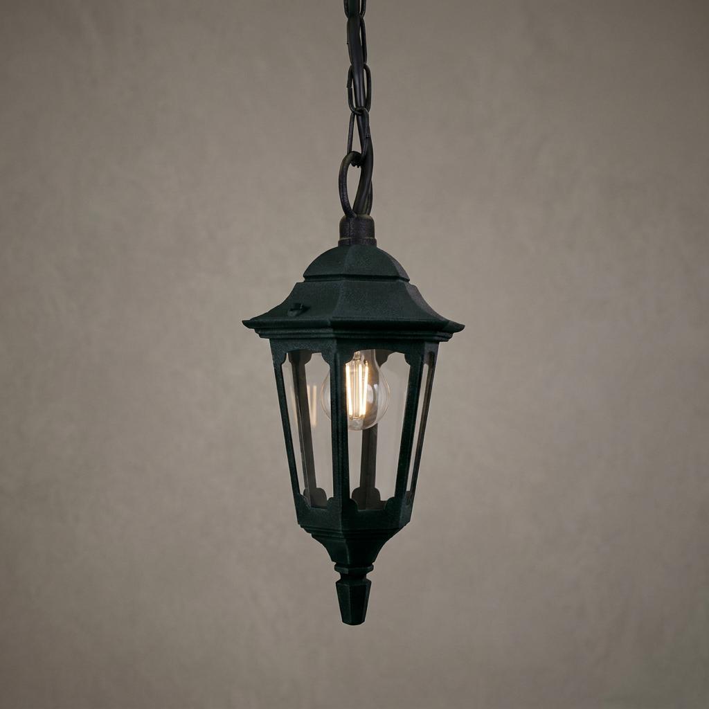 Parish black outdoor mini chain lantern with an illuminated bulb hanging against a plain grey textured wall.