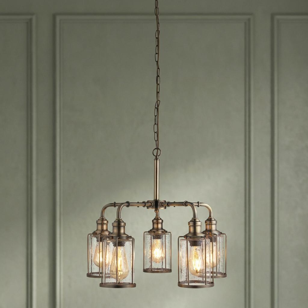 Industrial brass 5-light pendant with seeded glass shades and lit filament bulbs against a green paneled wall.