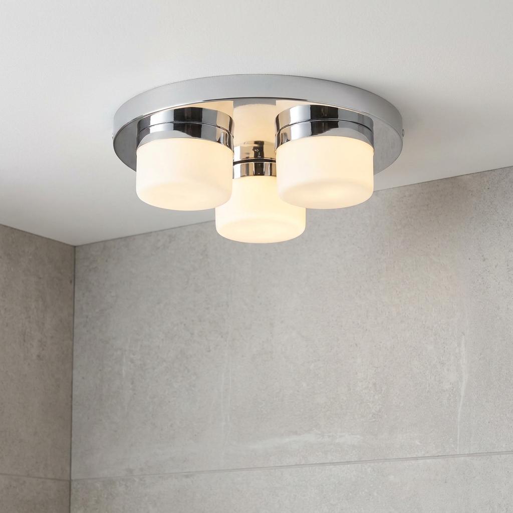 Three-light chrome flush mount bathroom ceiling fixture with opal glass shades against grey tiled walls.
