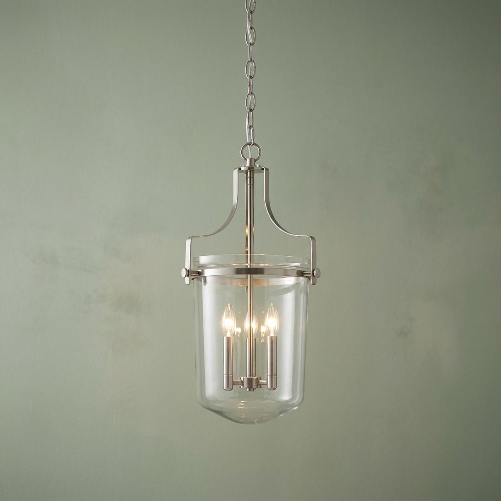 Three-light lantern pendant with a brushed nickel frame and clear glass shade against a sage green wall.