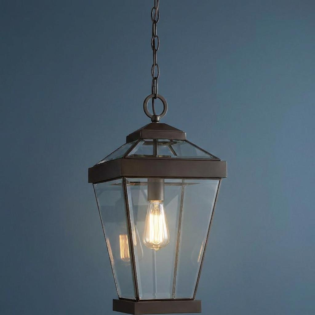 Bronze outdoor pendant lantern with clear glass panels and a lit Edison bulb against a blue wall.