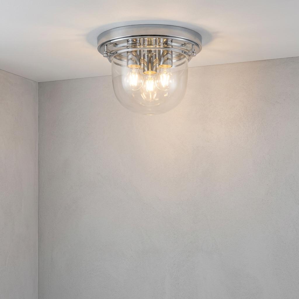 Chrome three-light flush mount ceiling fixture with a clear glass shade, illuminated in a grey room corner.