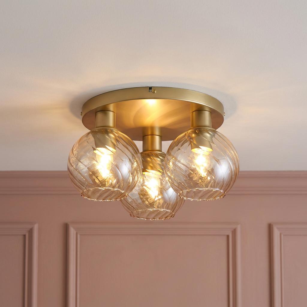A gold ceiling light with three glowing swirl glass globes mounted above a pink paneled wall.