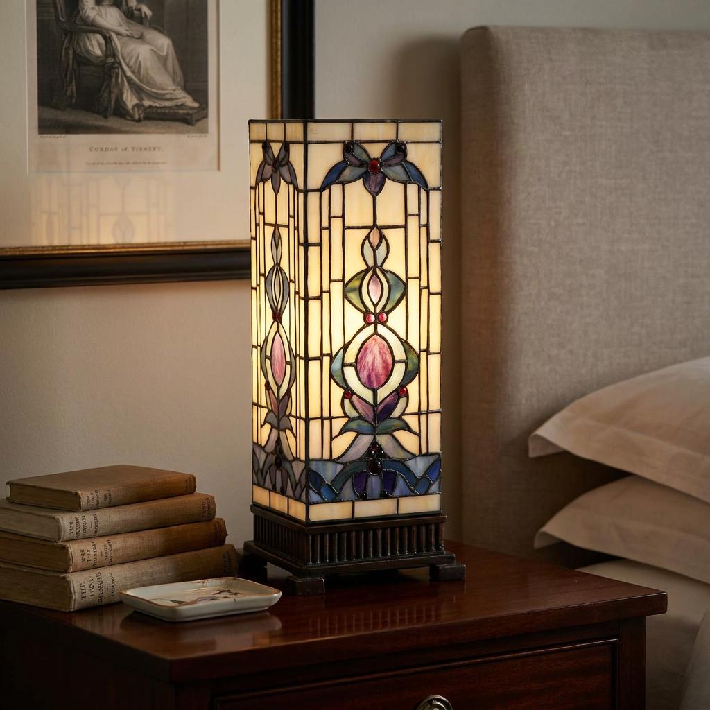 Lit rectangular Tiffany-style table lamp with floral stained glass on a wooden bedside table beside a stack of books.