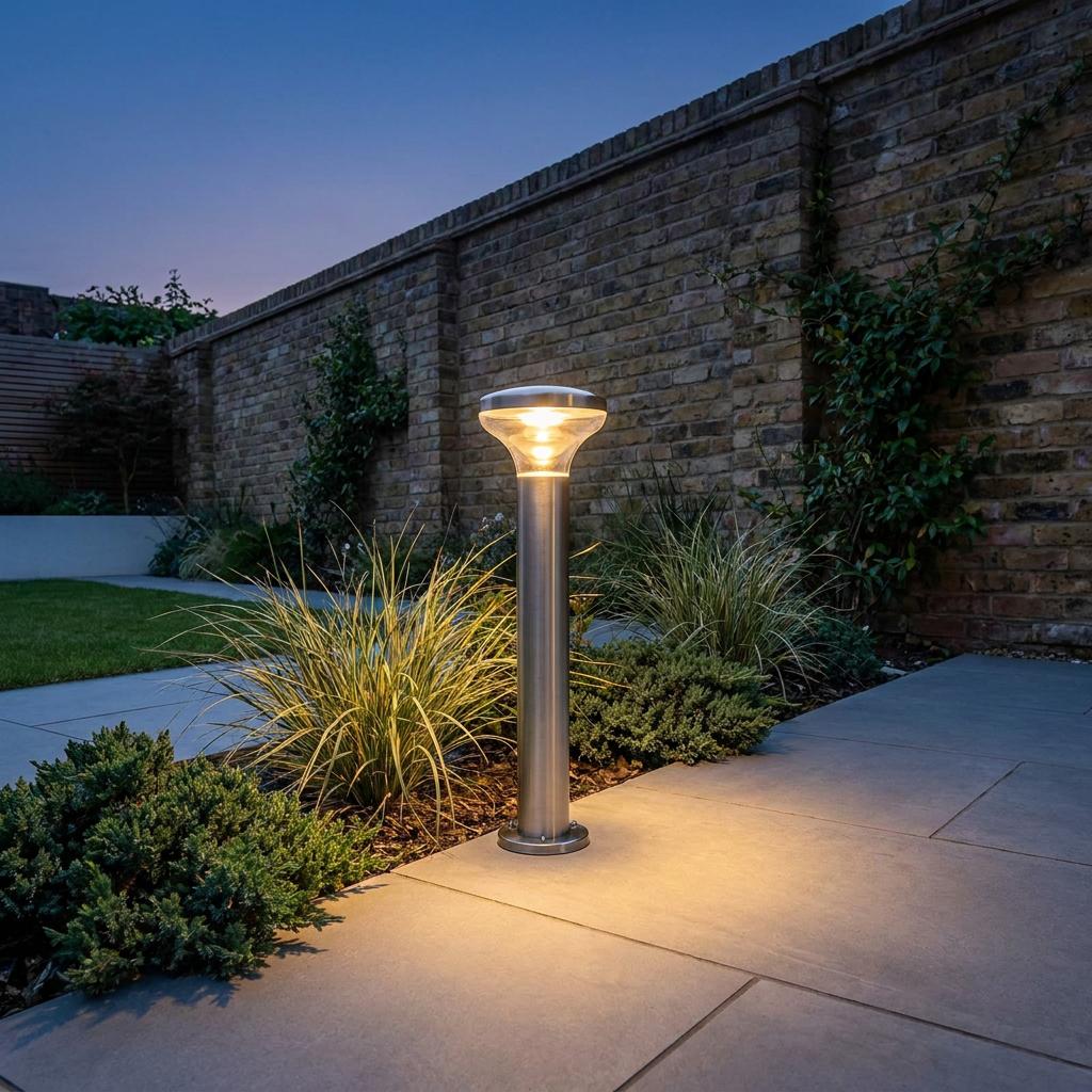 Brushed stainless steel outdoor bollard light illuminating a stone garden path at dusk against a brick wall.