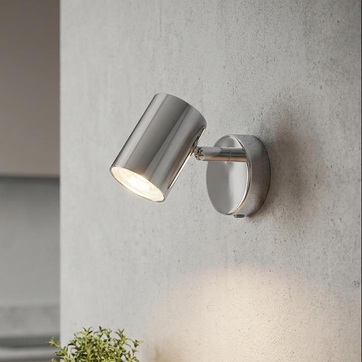 Chrome adjustable wall spotlight illuminated on a textured wall above a wooden shelf with a small plant.