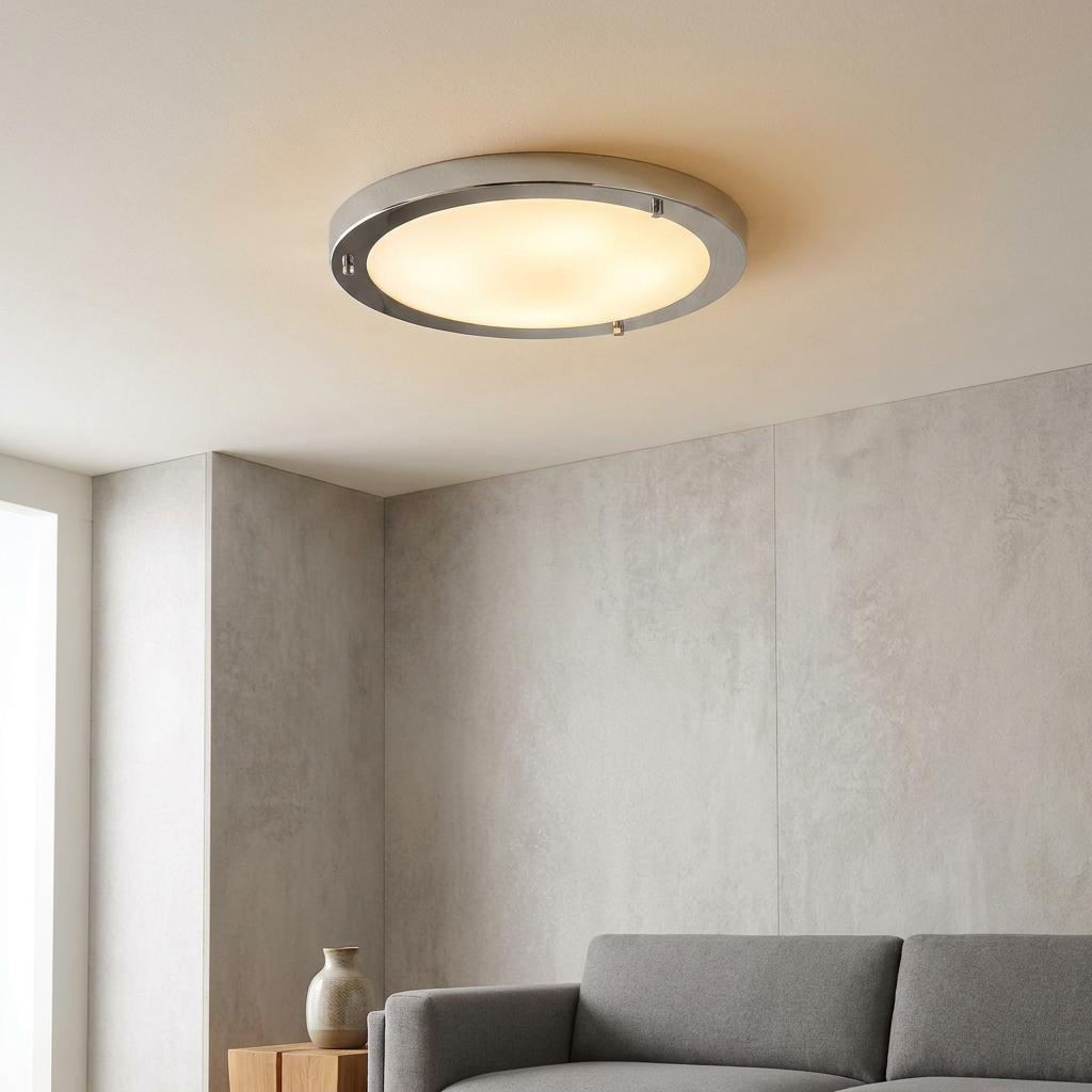 Circular chrome flush ceiling light with a frosted diffuser glowing above a grey sofa in a minimalist room.