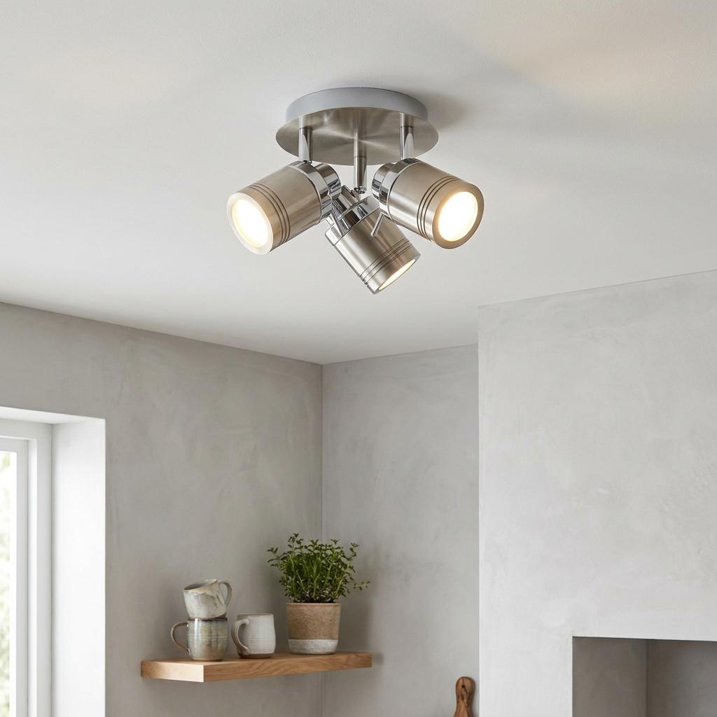 Three-light silver flush ceiling spotlight fixture illuminated above a wooden shelf in a modern interior.