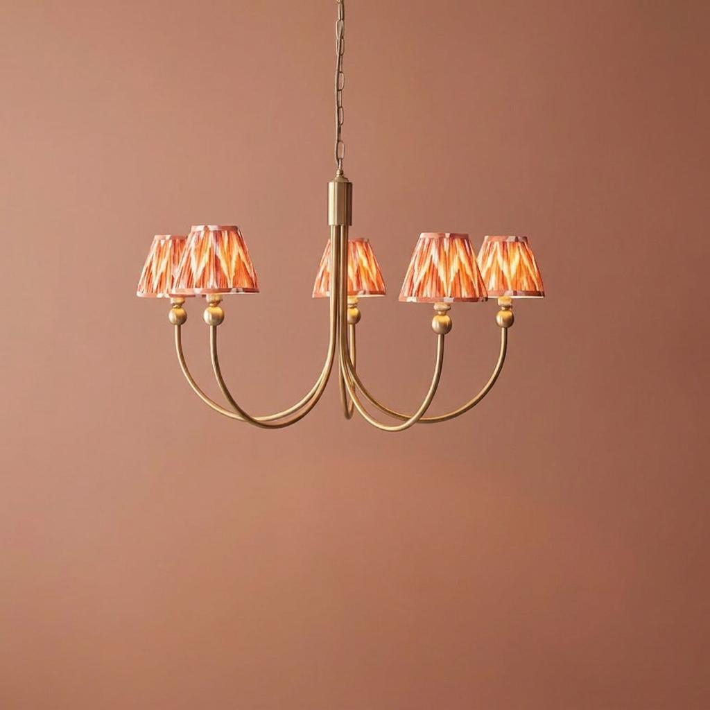 Santiago five-arm brass chandelier with orange zigzag pleated lampshades against a terracotta background.