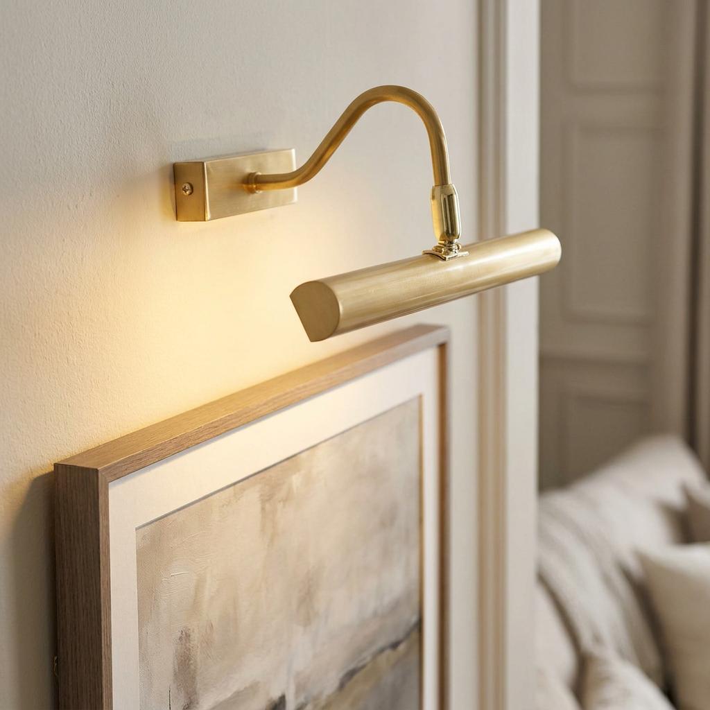 Satin brass picture light with a curved arm illuminating a framed painting on a neutral wall.