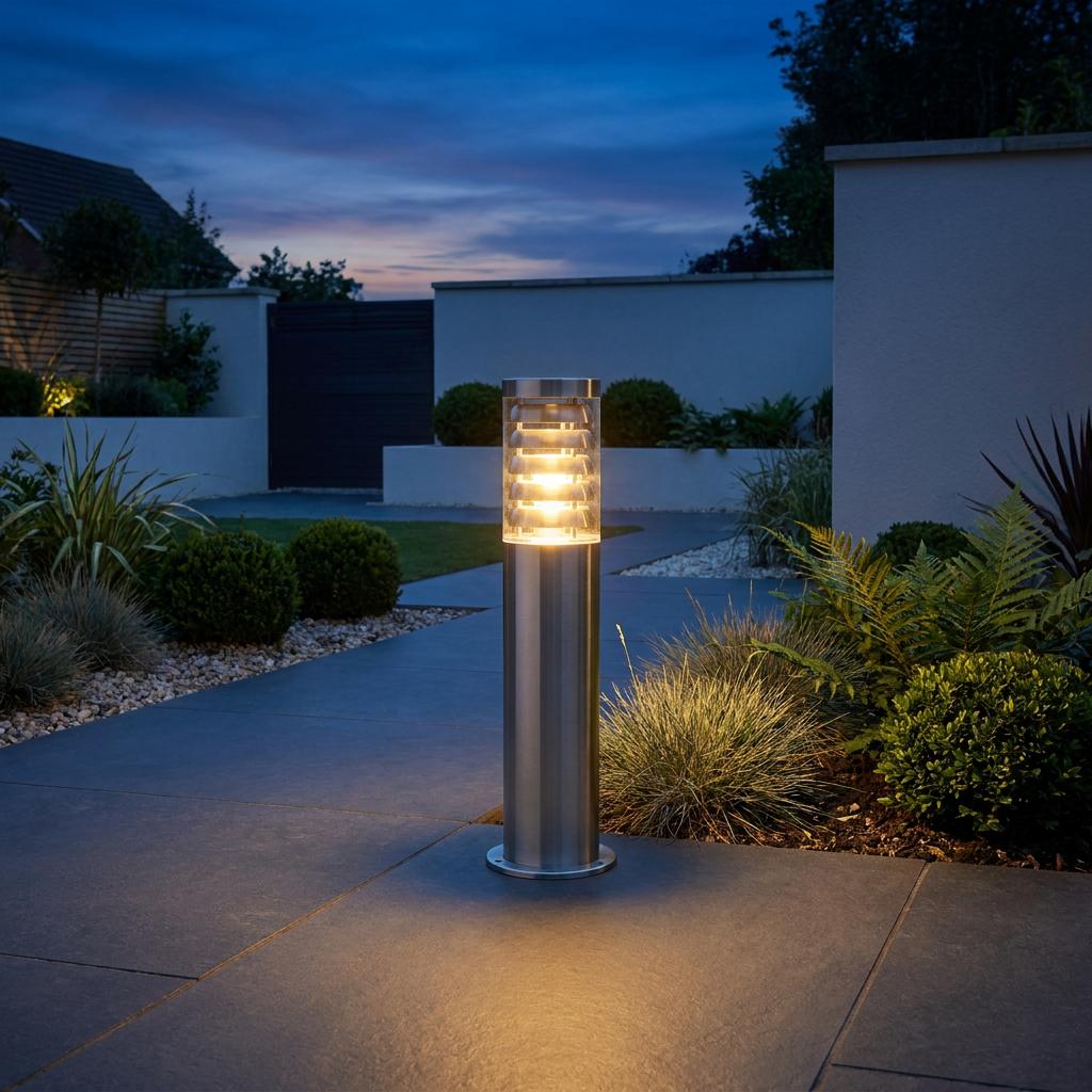 A brushed stainless steel outdoor bollard light illuminates a modern stone-paved garden path at twilight.