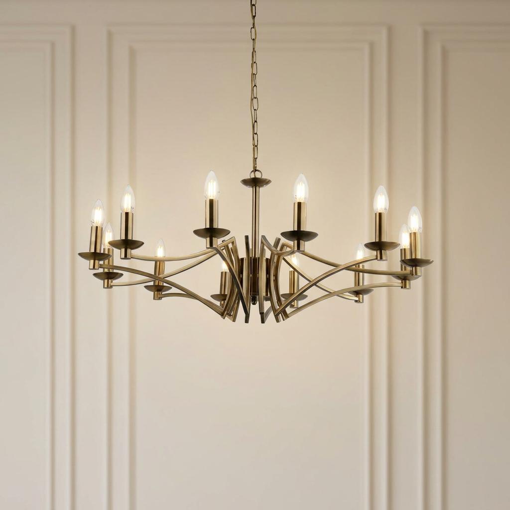 Searchlight Ascot 12-light antique brass chandelier with sweeping arms against a white paneled wall.