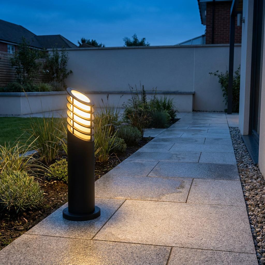 Modern black bollard path light with a slatted top glowing warm yellow on a paved garden walkway.