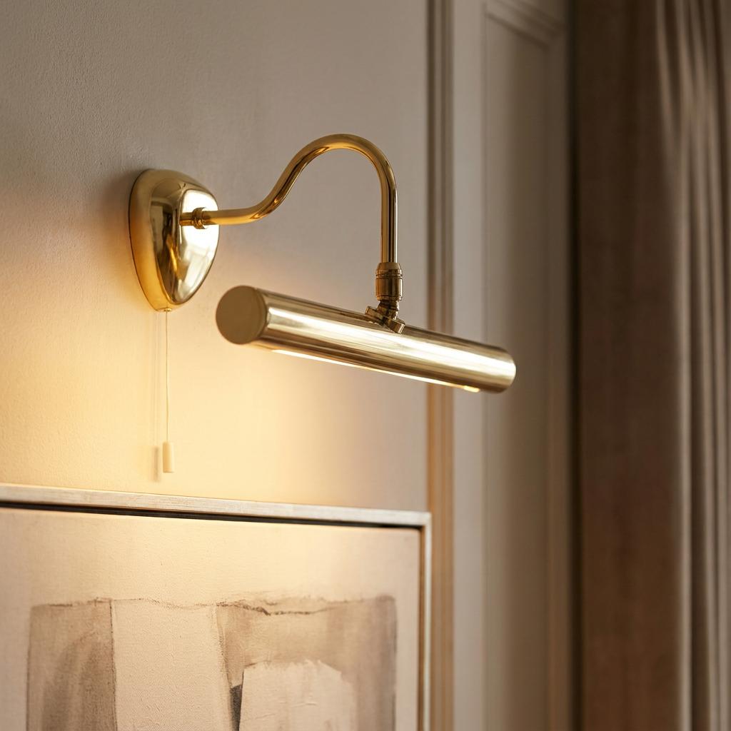 Polished brass picture light with a curved arm and pull cord illuminating a painting on a wall.